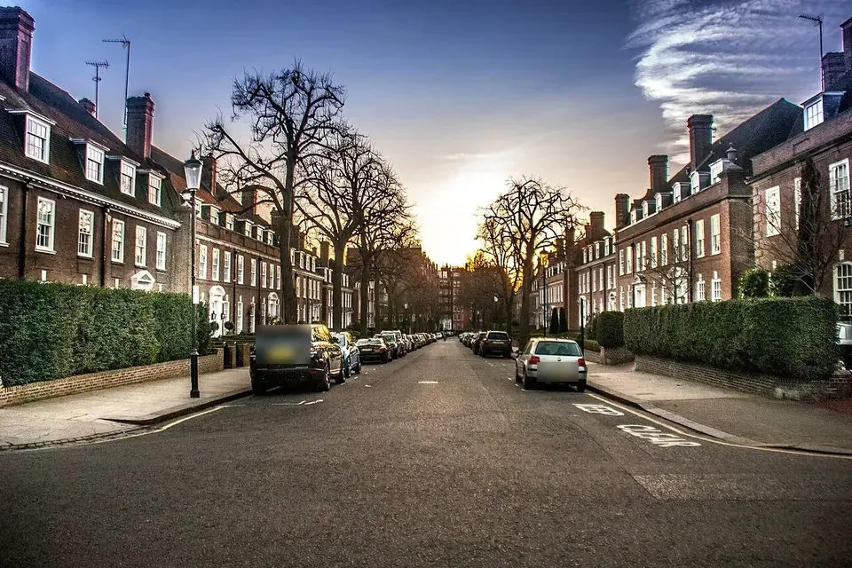 A quiet residential street lined with red-brick mansion blocks, hedges, bare plane trees and parked cars at sunset