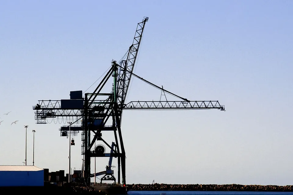 Silhouette of a ship-to-shore container crane with its boom lowered, against a pale blue sky over a harbour breakwater