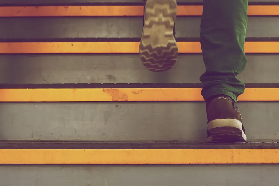 A person in green trousers and brown shoes climbing grey concrete stairs with bright yellow edges, seen from behind at step level