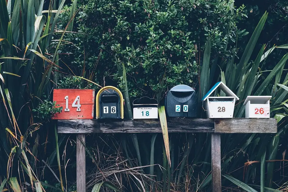 A row of six differently shaped and numbered mailboxes on a wooden stand in front of dense green foliage