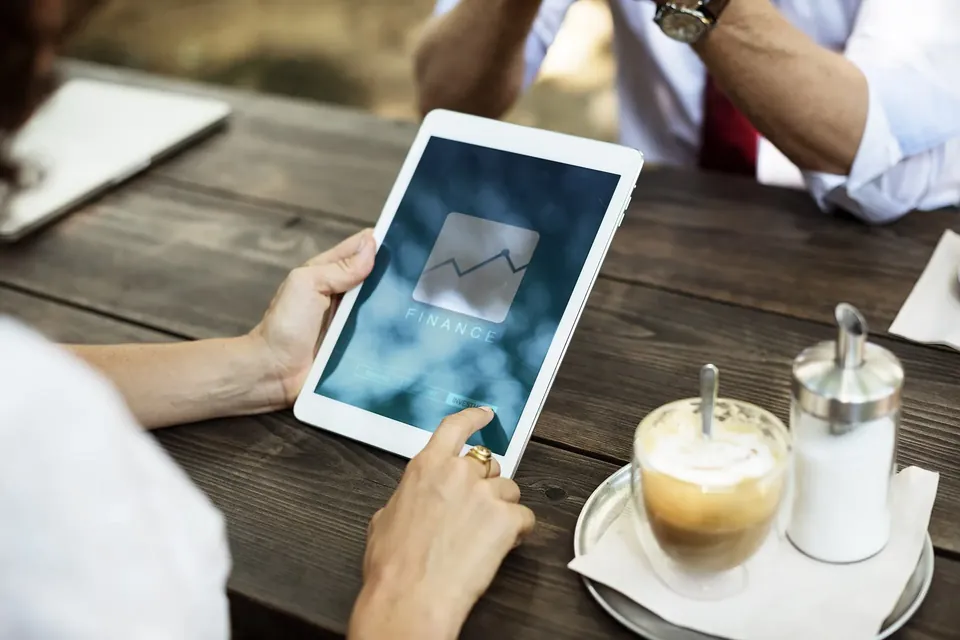 Hands holding a white tablet showing a simple finance chart icon, beside a glass of coffee on a wooden café table