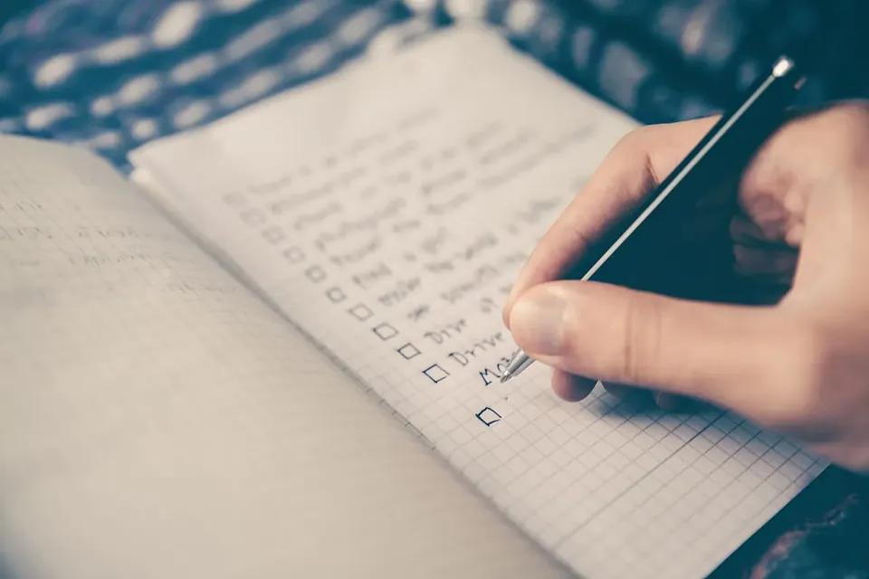 A hand holding a pen ticks boxes on a handwritten checklist in a squared notebook